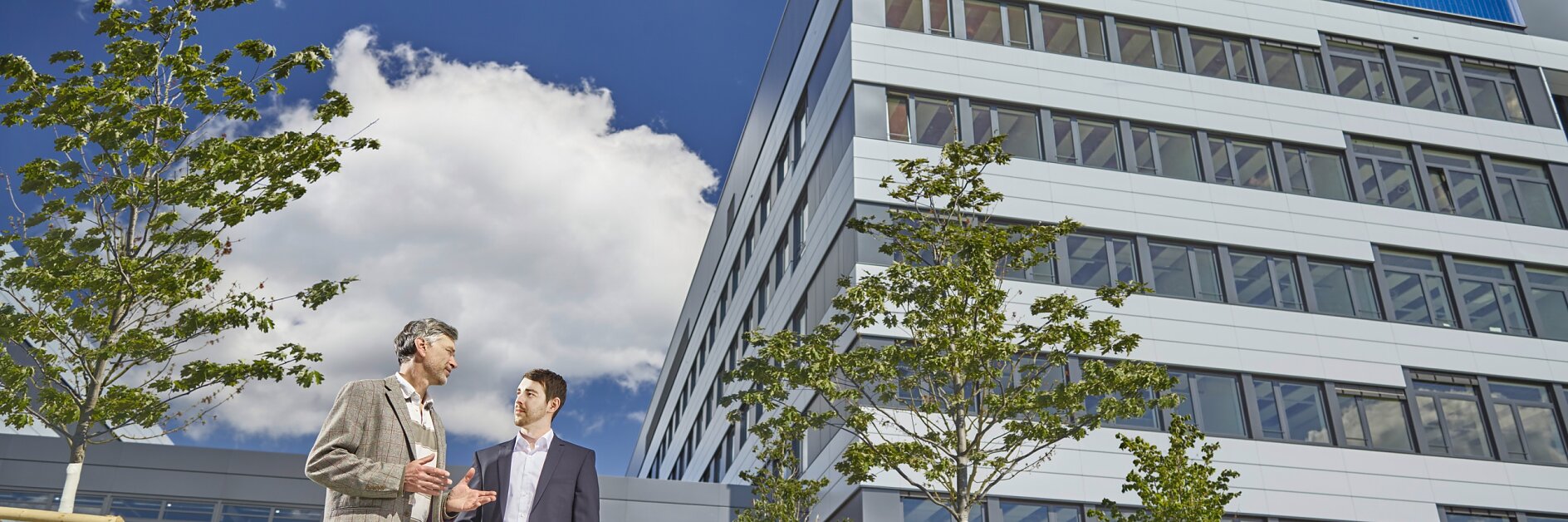 Two men in business attire stand conversing outside a modern multi-story building with the tesa tape logo. The sky is clear with a few clouds, and there are trees and greenery in the surroundings. (This text has been generated by AI)