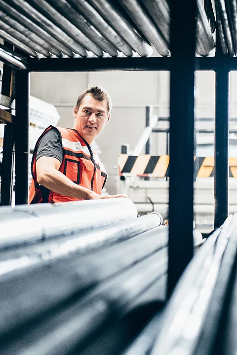 A person wearing an orange safety vest stands in a warehouse filled with metal pipes. They are looking to the side, surrounded by the stacked pipes on metal racks, with a truck partially visible in the background. (This text has been generated by AI)