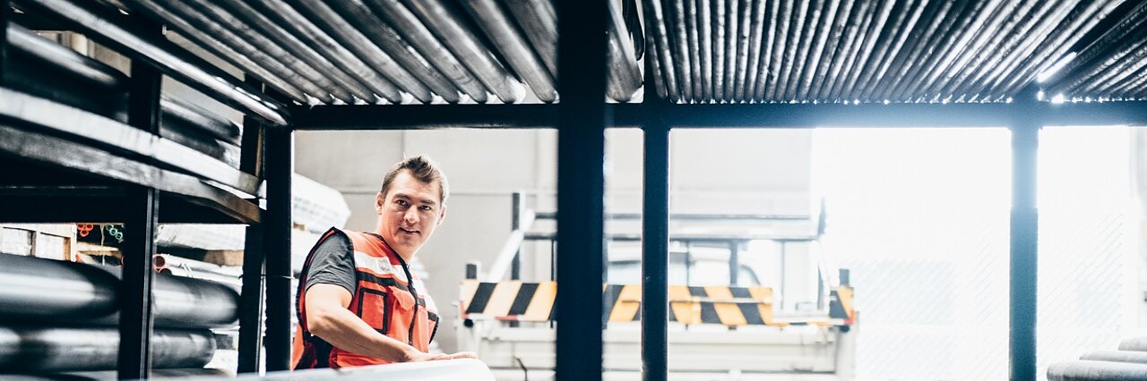 A person wearing an orange safety vest stands in a warehouse filled with metal pipes. They are looking to the side, surrounded by the stacked pipes on metal racks, with a truck partially visible in the background. (This text has been generated by AI)