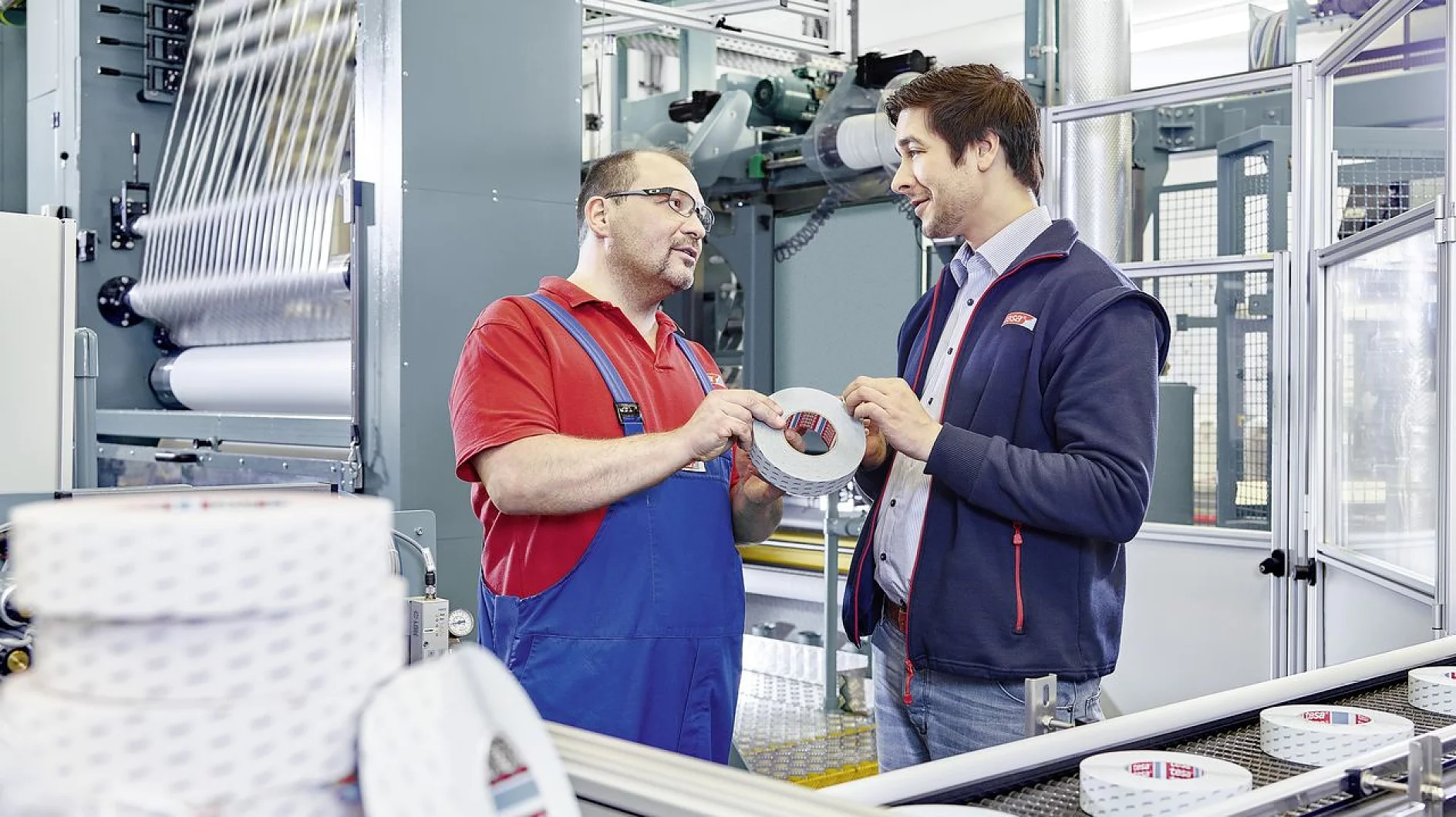 Two men in a manufacturing setting discuss while holding a roll of tesa tape. One wears a red polo and blue overalls, and the other wears a dark jacket and blue shirt. Machinery and conveyor belts are visible in the background. (This text has been generated by AI)