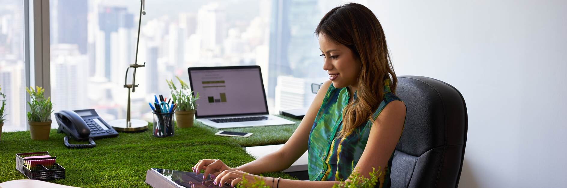 Concept of ecology and environment: Young business woman working in modern office with table covered of grass and plants. She types on tablet pc