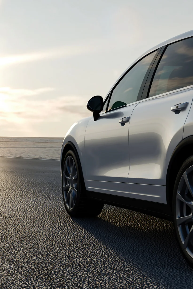 A white SUV is parked on a road near the ocean, with the sun low in the sky, creating a bright and reflective atmosphere. The car is positioned to the right, and the coastline is visible in the background. (This text has been generated by AI)