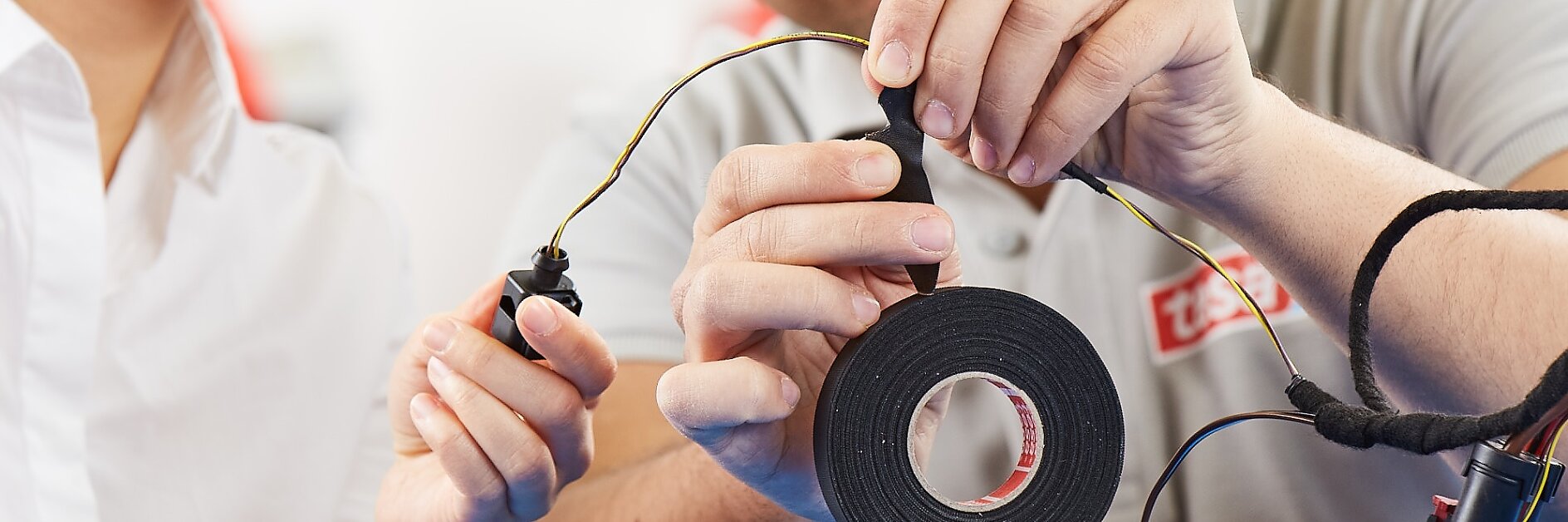 Two people are working together on an electronics project. One person holds a roll of black tesa tape, while both handle wires with black connectors. They are focused on assembling or repairing the components. (This text has been generated by AI)