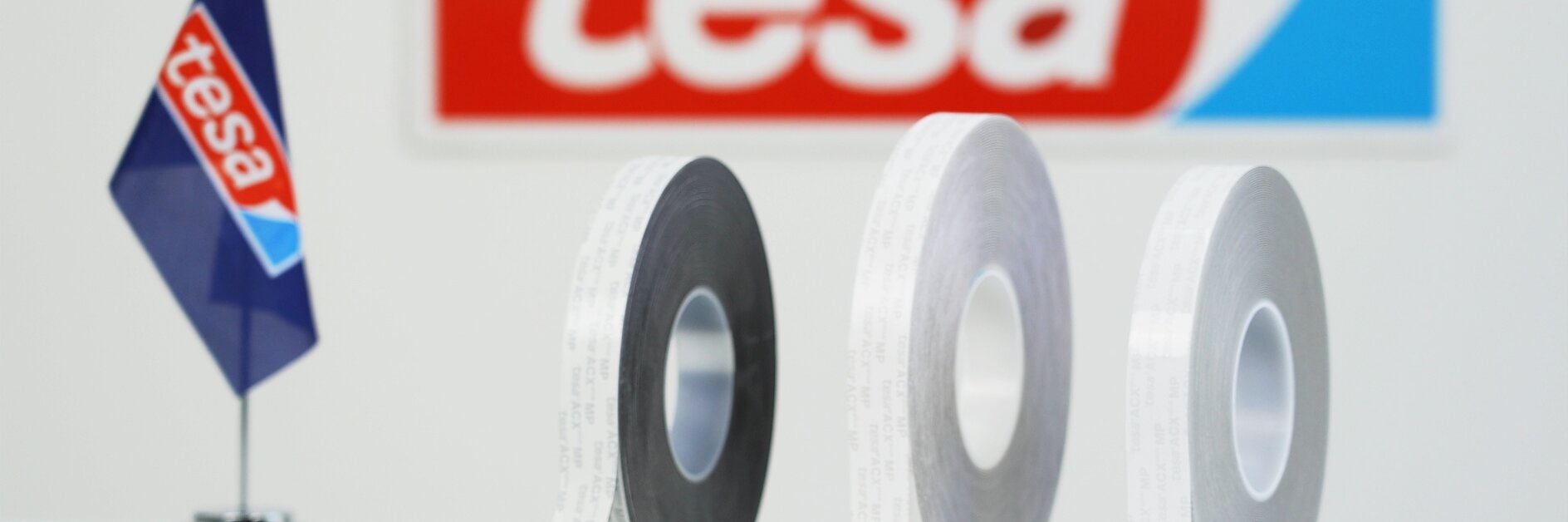 Three rolls of adhesive tesa tape in black, grey, and white are displayed on a surface. A small flag with the tesa logo is on the left, and a larger tesa sign is in the background. (This text has been generated by AI)