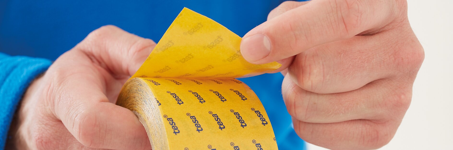 Close-up of hands holding and peeling away a piece of yellow adhesive tesa tape from a roll. The tesa tape has a repeated tesa logo printed on it. The person is wearing a blue long-sleeve shirt. (This text has been generated by AI)