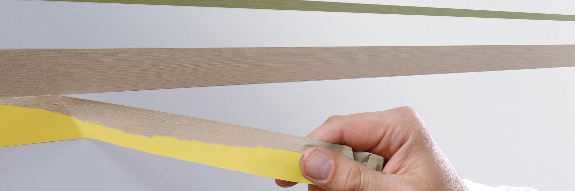 A person wearing a white sleeve removes yellow tesa tape from a wall. The wall has three painted horizontal stripes in green and two shades of brown. The tesa tape is being peeled off a light brown stripe. (This text has been generated by AI)