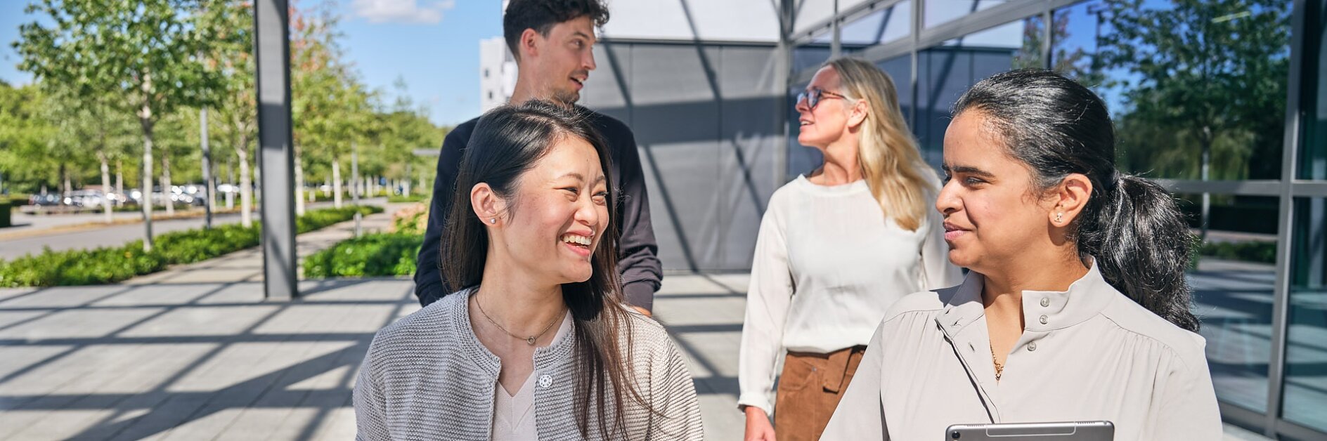 Four people walk and talk outside a modern building. Two women in the foreground are engaged in conversation, one holding a tablet secured with tesa tape. In the background, a man and a woman walk together. The area is paved and lined with trees. (This text has been generated by AI)