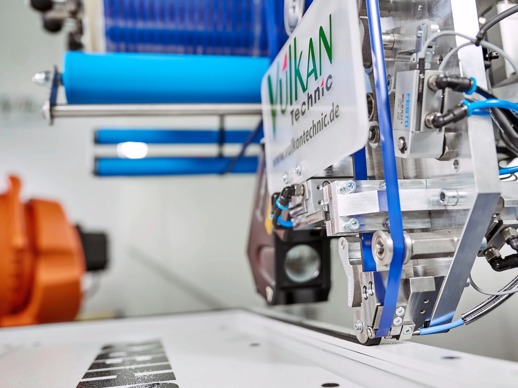 Close-up of industrial machinery in a manufacturing setting. The machine features metal components and blue tubing. Theres a spool of blue tesa tape material in the background, and a label on the machine reads “Mikan Technic.”. (This text has been generated by AI)