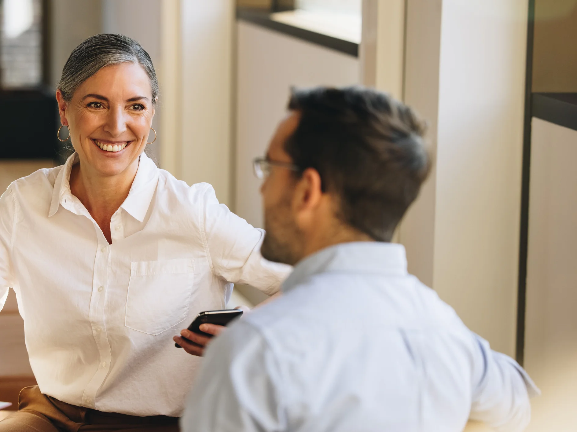 A woman with gray hair is smiling and holding a smartphone, sitting across from a man wearing glasses. Both are dressed in casual business attire and sitting in an office environment with large windows. (This text has been generated by AI)