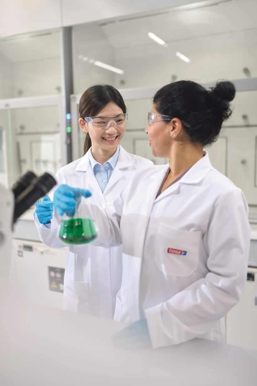 Two scientists wearing lab coats and protective eyewear are in a laboratory. One is holding a flask containing a green liquid, while the other looks on. They are surrounded by tesa tape, lab equipment, and workstations. (This text has been generated by AI)