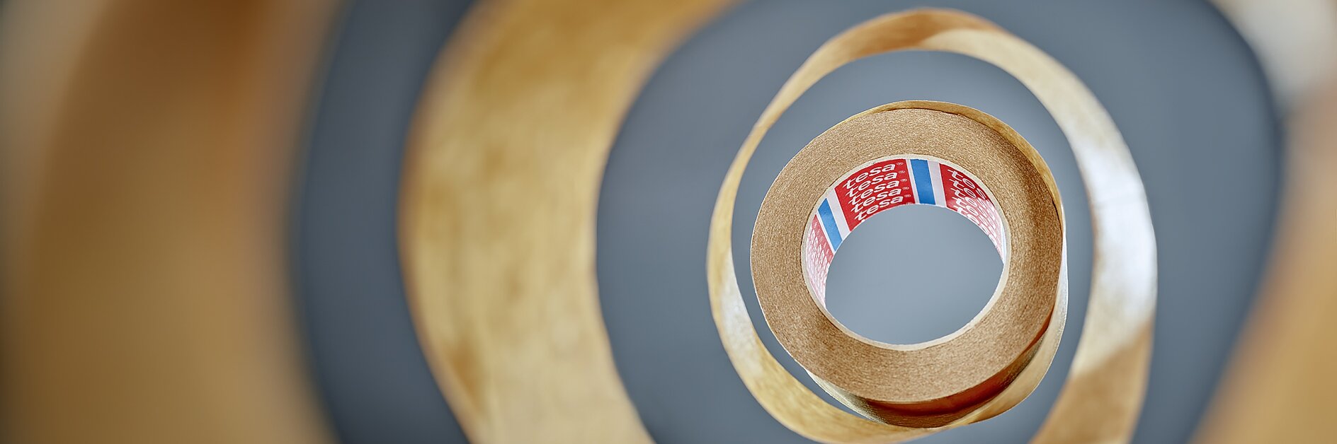 View from the center of a roll of tesa tape, creating a spiral pattern. The tesa tape is brown with a partially visible red label inside, featuring text and colored stripes. The background is blurred, emphasizing the layered effect of the tesa tape. (This text has been generated by AI)