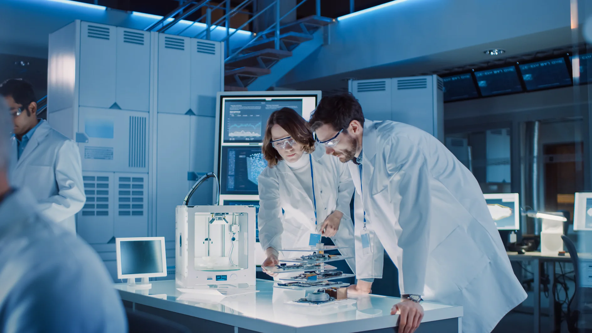 Diverse International Team of Industrial Scientists and Engineers Wearing White Coats Working on Heavy Machinery Design in Research Laboratory. Professionals Using 3D Printer, Computers and Microscope